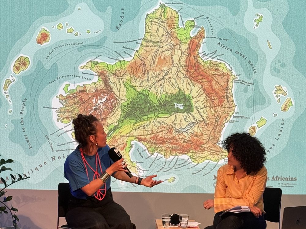 Elodie Rama et Zoubida Debbagh assises discutant posant devant un visuel visioprojeté de la carte Maison des Mondes Africains