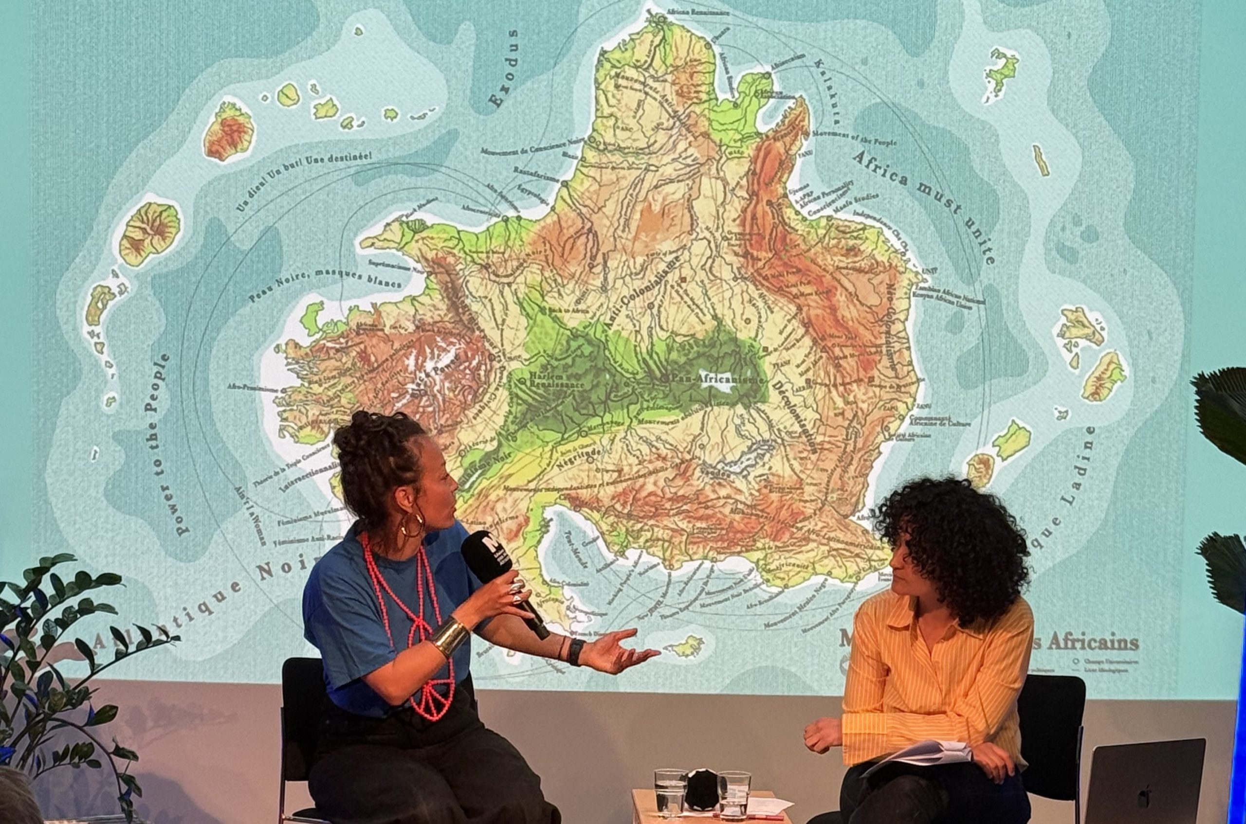 Elodie Rama et Zoubida Debbagh assises discutant posant devant un visuel visioprojeté de la carte Maison des Mondes Africains